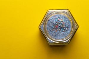 Opened glass jar with blue powder and multi-colored granules, on a yellow background. Copy space. Loose colored paint. Top view. photo