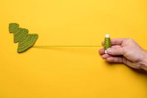 Knitted Christmas tree - a symbol of the New Year and a hand unwinding a spool of thread, on a yellow background. Copy space. photo