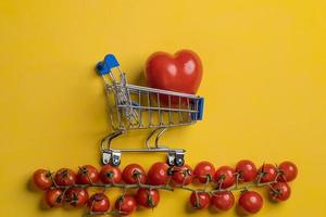 Red tomato in the form of a heart in a grocery trolley, next to cherry tomatoes, on a yellow background. Healthy food concept. photo