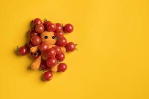 Cute homemade knitted toy man in red cherry tomatoes, on a yellow background. Copy space. Top view. photo