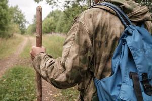 Traveler with a stick, in camouflage clothes and with a backpack stands against the backdrop of a forest road and trees. Active lifestyle. photo