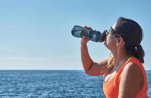 Latin woman,resting regaining strength, eating, drinking water, after a gym session, wearing orange top, burning calories keeping fit by the sea and smart watch photo