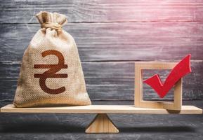 Red vote tick and a ukrainian hryvnia money bag on scales. Estimating cost of making a decision and consequences in the future. Corruption risks. Concept of lobbying for decisions and laws. photo