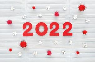 números de fieltro rojo 2022 del año sobre fondo de máscaras médicas azul claro. copos de nieve blancos de madera y pompones rojos alrededor como molécula de coronavirus. revisión de los resultados del año de la pandemia de covid-19. foto