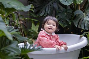 Caucasian little toddler baby girl smiling and playing inside the bathtub surrounding by tropical plant for better air purifying and sustainable home design concept photo