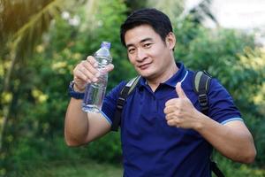 Handsome Asian man traveler holds bottle of drinking water to drink outdoors. Concept , Drinking water for health, Healthy lifestyle.Quenching thirst, reduce fatigue, refreshes body. photo