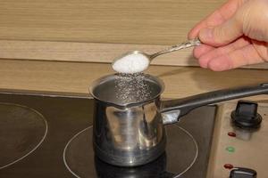 hand putting sugar with spoon in empty pot on stove photo