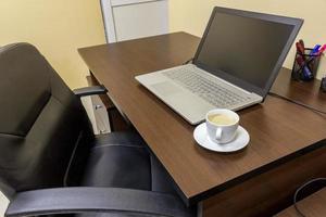 cómodo lugar de trabajo en la oficina con mesa de madera, silla de cuero negro, computadora portátil y taza de café. foto