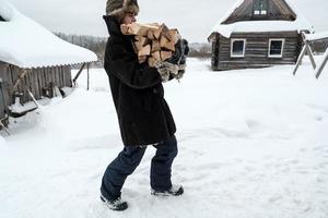 Man, in warm clothes, carries an armful of firewood, in a rural yard, on a winter evening. Heating season in the village. photo