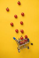 Overturned decorative grocery cart and cherry tomatoes that fell out of it, a fallen wheel next to it, against a yellow background. Top view. photo
