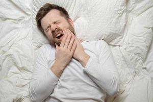 Man suffers from unbearable toothache, he holds hands on cheek and groans in pain, closing eyes and lying in bed. Top view. Close-up. photo