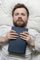 Stunned man lies in bed clutching the book he has finished to chest and looks up enthusiastically. Top view. photo