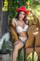 sensual brunette woman with sexy country look on chair. Portrait of a girl with white lingerie and red hat. Girl  having fun with in  brown leather boots at the ranch. American sexy country style photo