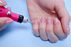 el proceso de hacer una hermosa manicura en los dedos de un dedo procesando una uña con una máquina especial en un salón de belleza de uñas sobre un fondo azul foto