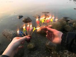 Burning happy birthday inscription made of holiday candles in the hands of a man and a woman opposite the water of the ocean lake river. Concept birthday celebration in nature, outdoors photo