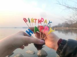 inscripción de feliz cumpleaños hecha de velas navideñas en manos de un hombre y una mujer frente al agua del río del lago oceánico foto