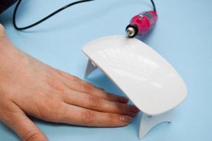 The process of making a beautiful manicure on the hands on the fingers with the help of a special machine and a lamp for drying the varnish in the nail beauty salon on a blue background photo