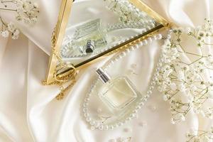 A chic perfume bottle and pearl beads are reflected in an elegant mirror on a beige satin fabric background. aroma presentation. photo