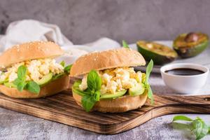 Delicious sandwich with scrambled eggs, avocado and basil in a bun on a board. Healthy food. photo