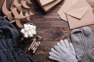 Winter still life. Marshmallow drink, warm clothes, craft envelopes and Christmas trees on a wooden photo