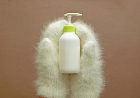 Cosmetic pump bottle of moisturizing lotion hold hands in mittens on pastel background. White corrugated bottle surface soap dispenser pump. Hand skin care lotion in winter. photo