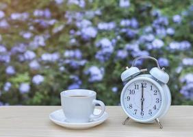 white vintage alarm clock and cup of coffee on the table in the garden with purple flowers background. photo