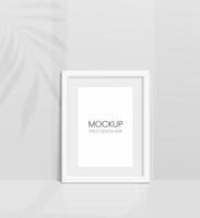 Realistic vector mockup with white photo frame. Shadow on the wall and empty place for your design. 3d soft light and overlay shadow from leaves of plant and window. Vertical poster or painting.