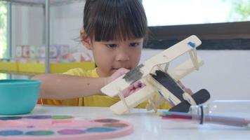 Porträt eines glücklichen asiatischen Mädchens mit einer Pinselmalerei auf einem hölzernen Spielzeugflugzeug im Klassenzimmer. Kunsthandwerk für Kinder. kreativer kleiner künstler bei der arbeit. video