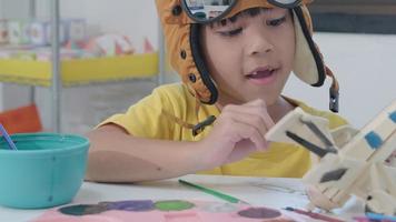 Porträt eines glücklichen asiatischen Mädchens mit einer Pinselmalerei auf einem hölzernen Spielzeugflugzeug im Klassenzimmer. Kunsthandwerk für Kinder. kreativer kleiner künstler bei der arbeit. video