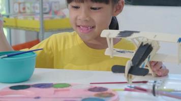 retrato de uma menina asiática feliz com uma pintura de pincel em um avião de brinquedo de madeira na sala de aula. artes e ofícios para crianças. pequeno artista criativo no trabalho. video