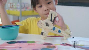 Portrait of a happy Asian girl with a brush painting on a wooden toy plane in the classroom. Arts and crafts for kids. Creative little artist at work. video