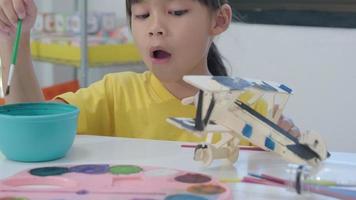 retrato de una chica asiática feliz con un pincel pintando en un avión de juguete de madera en el aula. artes y manualidades para niños. pequeño artista creativo en el trabajo. video
