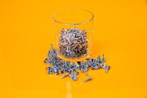 Many metal self-tapping screws made of steel in a glass, in bucket jar. self-tapping screw for metal, for iron, chrome-plated self-tapping screw, on an orange yellow background, photo