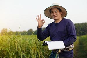 Asian man farmer is at paddy field, holds notebook paper to check growth and diseases of rice plants, make OK hand sign. Concept, Agriculture research and study to develop crop photo