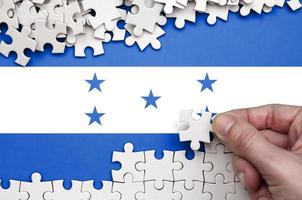 Honduras flag is depicted on a table on which the human hand folds a puzzle of white color photo