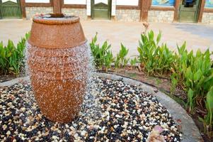 A beautiful little fountain in the form of a brown vase, a jug with falling drops of water on colored stones standing in a flower bed with green plants photo