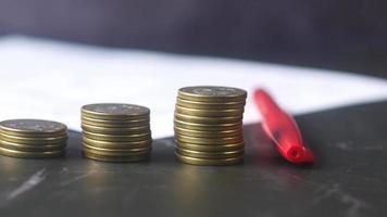 Close up of stacks of coins at different levels next to red pen and paper video