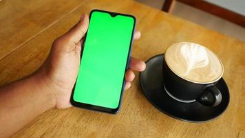 Adult holds phone with green screen at cafe table next to a latte in a mug video