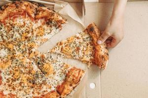 delivery de pizza. pizza italiana mano sostiene una rebanada de pizza. entrega de alimentos en línea. comida rapida en la mesa foto