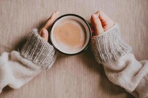 hands in a sweater hold hot cocoa, in a red mug, top view. A cozy photo with a mug in hand with copy space