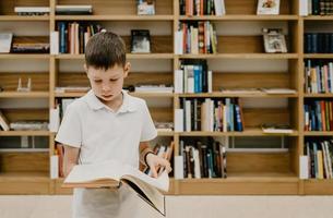 A boy stands in the library and reads a book while standing. Preparing for homework. The boy loves to read. Free space at the school. Extracurricular learning. photo