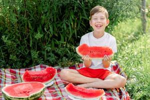 boy eating watermelon white t-shirt. Picnic with watermelons. The boy is holding a large piece of watermelon in his hands. Bright red juicy watermelon. photo