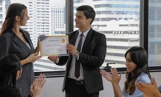 Young caucasian manager in suit giving mock up certificate to woman staff with happy smiling face while others applauding for congratulation in office meeting room. Selective focus. photo