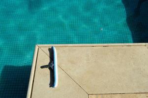 A blue brush with a sponge for cleaning the cleaning of the pool tending the tiles on the background of the pool with shallow square ceramic tiles and clear water photo