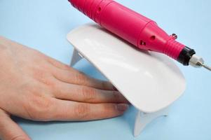 The process of making a beautiful manicure on the hands on the fingers with the help of a special machine and a lamp for drying the varnish in the nail beauty salon on a blue background photo