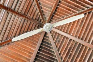 The texture of a brown wooden roof is abstract of the beams of logs arranged vertically horizontally and a large ceiling fan from the heat. The background photo