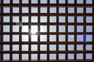 Texture of a brown wooden abstract lattice with square cells with holes of boards of log beams arranged vertically horizontally against the background of the sun and sky. The background photo