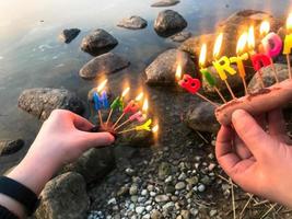 inscripción de feliz cumpleaños en llamas hecha de velas navideñas en manos de un hombre y una mujer frente al agua del río del lago oceánico. concepto de celebración de cumpleaños en la naturaleza, al aire libre foto