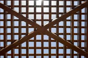 Texture of a brown wooden abstract lattice with square cells with holes from boards of log beams arranged vertically horizontally diagonally against the background of the sun and sky. The background photo