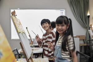 Portrait of Asian kids looking at camera and smiling with acrylic color picture painted on canvas in art classroom and creative learning with talents and skill at elementary school studio education. photo